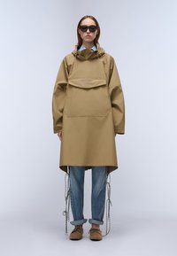 Anorak bej rezistent la apă, cu glugă, buzunar frontal și șnur ajustabil. Pantaloni blugi albastru deschis cu manșete rulate și pantofi maro.