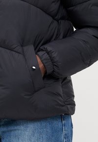 Svart pufferjakke med kviltet design, elastiske mansjetter og lomme med trykknapp. Materialet ser glatt og isolert ut.
