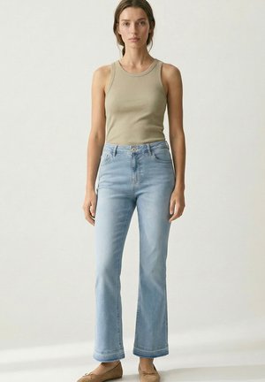 Woman stands facing forward wearing a beige sleeveless top, light blue high-waisted jeans, and tan flats against a plain background.