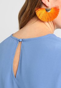 Blouse bleue avec un détail en forme de trou de serrure au dos, fermée par un bouton, associée à de grandes boucles d'oreilles en cuir orange et argent, présentant un design texturé.