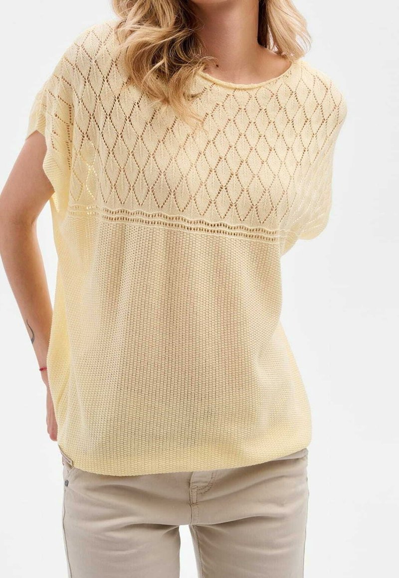 Femme portant un haut en maille jaune clair à manches courtes avec des motifs losanges et gaufres et un pantalon beige sur fond blanc.