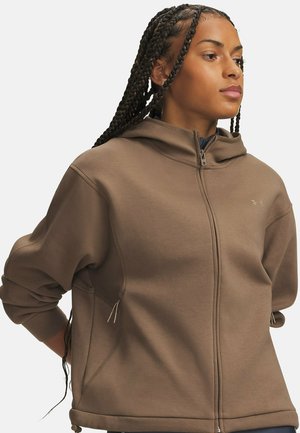 Vrouw met lange vlechten draagt een bruine hoodie met rits, staat met de handen achter de rug en kijkt opzij tegen een effen achtergrond.