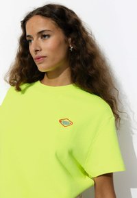 T-shirt en coton jaune fluo à manches courtes, orné d'un petit patch coloré en forme de diamant portant l'inscription "Sunset Club." Texture lisse.