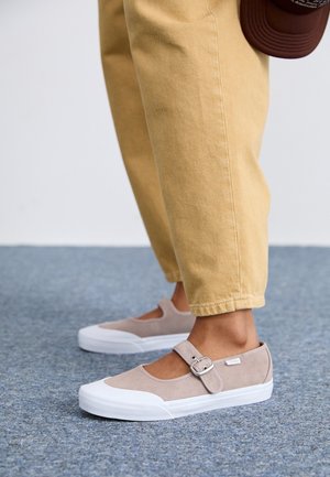 Stopy w beżowych zamszowych trampkach Mary Jane z białymi gumowymi podeszwami i paskami na klamrę, w połączeniu z jasnobrązowymi spodniami na niebieskim dywanie.