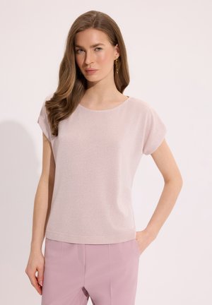 Młoda kobieta z długimi, falowanymi włosami, ubrana w jasnoróżowy krótki top z krótkim rękawem i liliowe spodnie, pozująca z jedną ręką w kieszeni na białym tle.