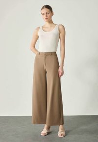 Calças largas bege com cintura alta, feitas de tecido leve. Combinadas com uma blusa justa branca e sandálias claras.