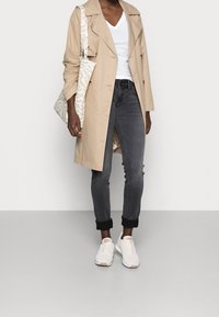 Trench coat beige, camiseta blanca de escote en V, jeans oscuros con dobladillo, zapatillas blancas y una bolsa tote con un diseño en crema y beige.
