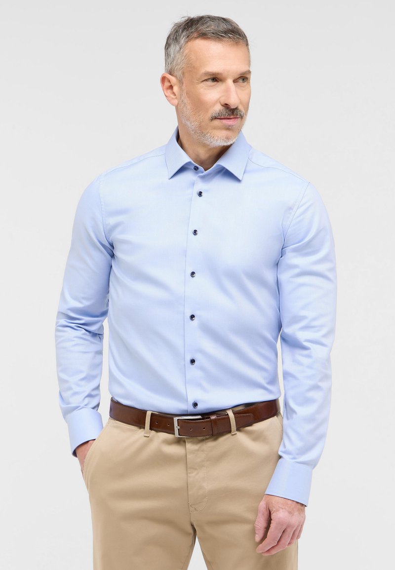 Lichtblauw langarmshirt met een button-down kraag, voorzien van donkere knopen, gecombineerd met beige broek en een bruine riem.