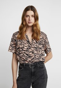 Camisa de manga corta de tela rosa claro, con un patrón negro de hojas, dos bolsillos en el pecho y un escote en V. Combinada con un cinturón negro con tachuelas.