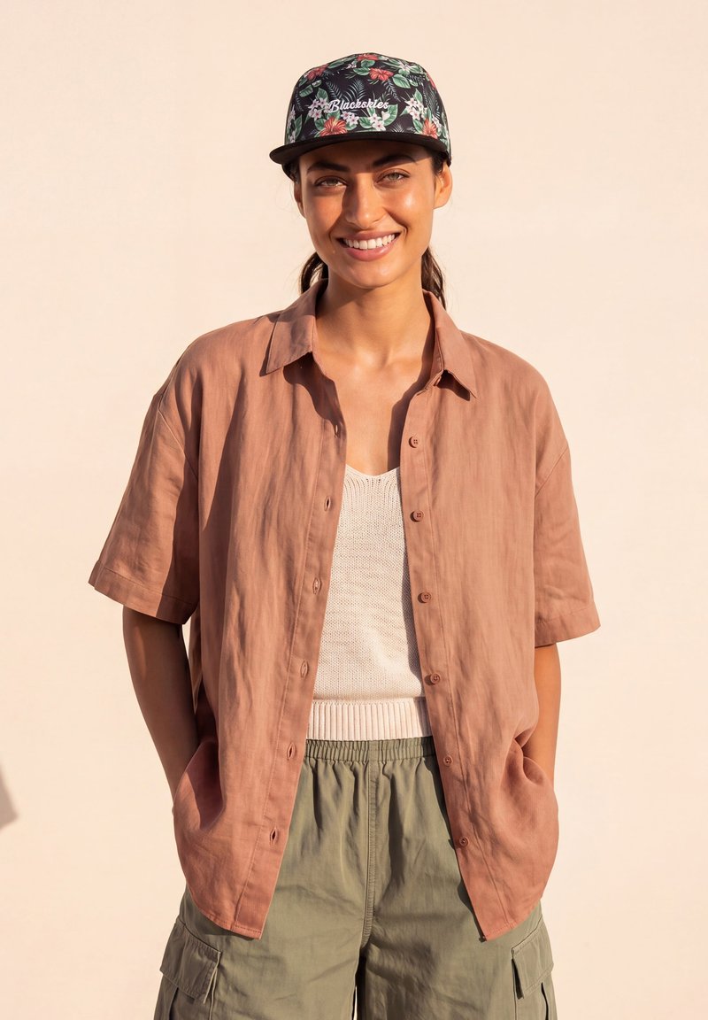 Mujer sonriente vistiendo una gorra con flores, camisa marrón suelta con botones, top de punto blanco y pantalones cargo verdes con las manos en los bolsillos.