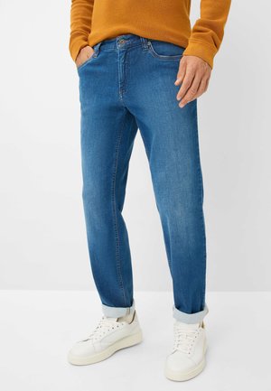 Jean en denim bleu avec une coupe slim, dotés de poches avant et de manches retroussées, associés à des baskets blanches et un pull en laine texturé de couleur moutarde.