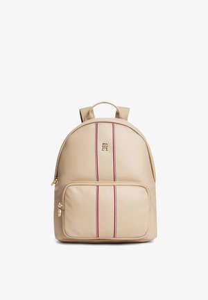 Sac à dos beige avec poche zippée avant, double rayure verticale rouge et blanche, logo doré et bretelles réglables.