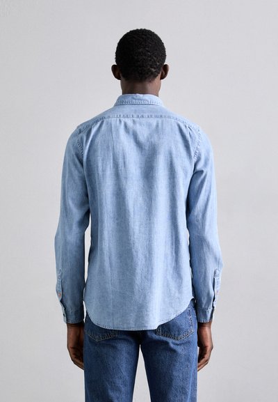 Camisa de mezclilla azul claro con cuello abotonado, mangas largas y dobladillo curvado. Presenta detalles de costura y puños con botones.