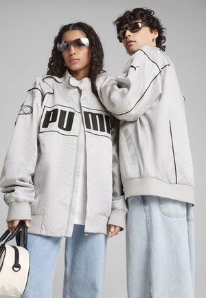Veste grise surdimensionnée Puma avec des accents noirs, arborant un grand logo, des poignets côtelés et une texture lisse et rembourrée, associée à un jean bleu clair.
