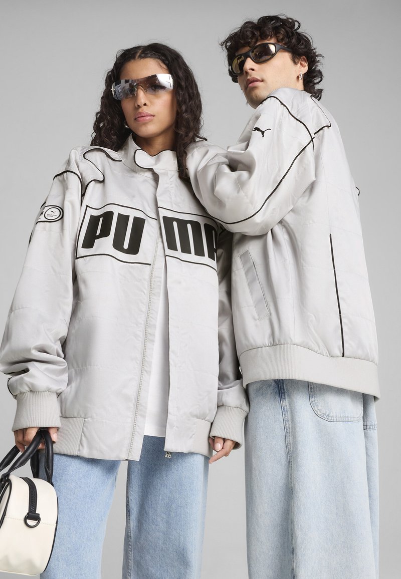 Puma oversized grå jakke med sorte akcenter, med en fremtrædende logo, ribbede manchetter og en glat, polstret tekstur, parret med lyseblå jeans.