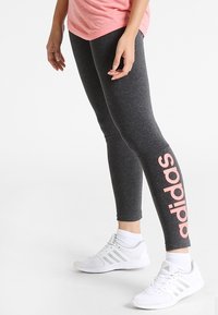 Szare legginsy z różowym logo "adidas" po boku, wykonane z miękkiego, elastycznego materiału; zestawione z białymi butami sportowymi.