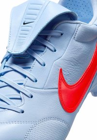 Világoskék mesterséges foci cipő piros Nike pipa emblémával, texturált bőrből készült, perforált díszítésekkel. Lapos nyelvével és fűzős záródásával rendelkezik.