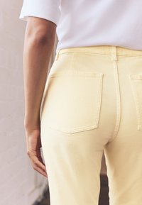 Pantalon en jean jaune avec poches arrière, taille haute et texture lisse. Le bras du modèle est placé à côté du pantalon.