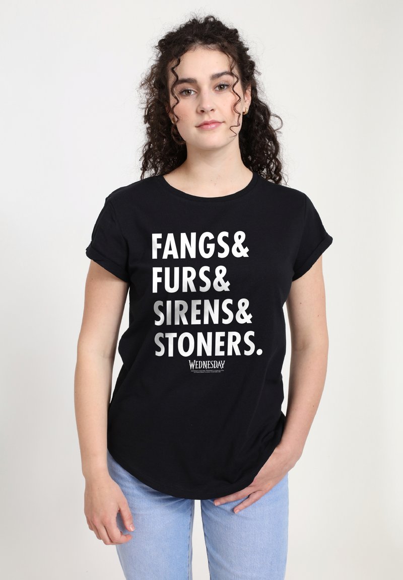 Mladý člověk s kudrnatými vlasy má na sobě černé tričko s tučným bílým nápisem "Fangs& Furs& Sirens& Stoners." a světle modré džíny.