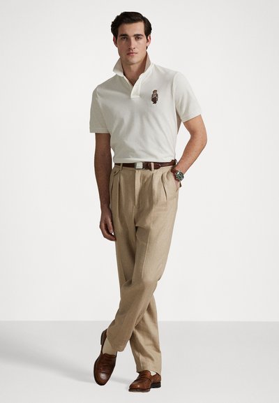 Tricou polo alb cu logo brodat cu un urs, pantaloni plisati bej, pantofi din piele maro și curea maro. Modelul stă pe un fundal deschis.