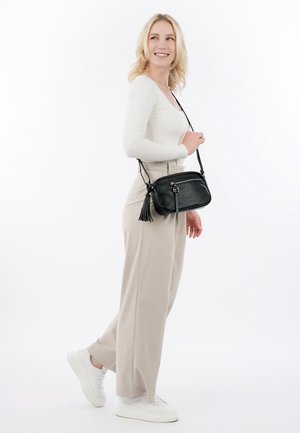 Blonde Frau trägt weißes Oberteil und beige Hose, steht lächelnd da und trägt eine schwarze Umhängetasche mit Quasten-Detail.