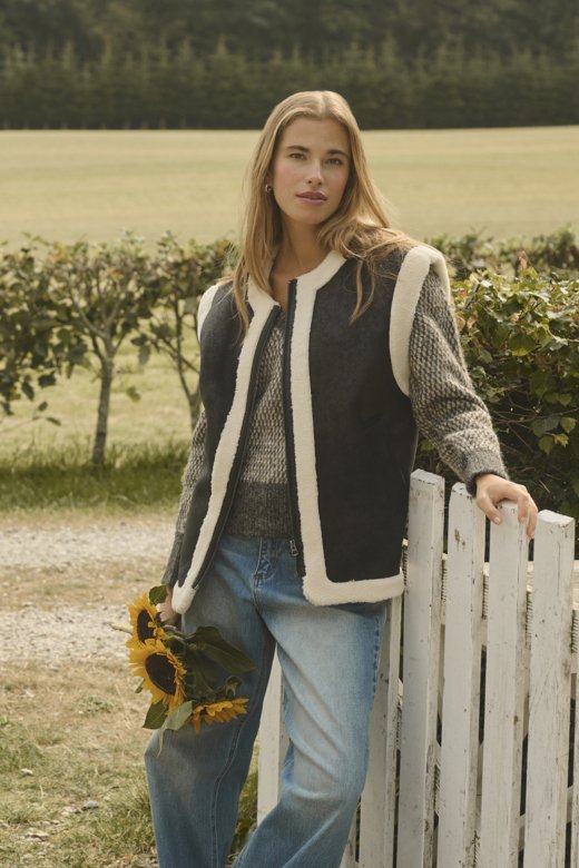 Žena u krznenom prsluku i pletenom džemperu drži suncokrete, naslonjena na bijelu drvenu ogradu s pozadinom zelene površine i polja.