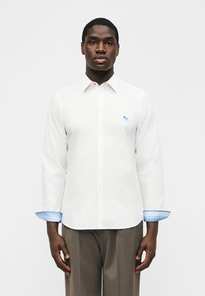 Mand iført en hvid, langærmet skjorte med knapper, med blå broderet zebra-logo og blåstribede indvendige manchetter, stående mod en ensfarvet baggrund.