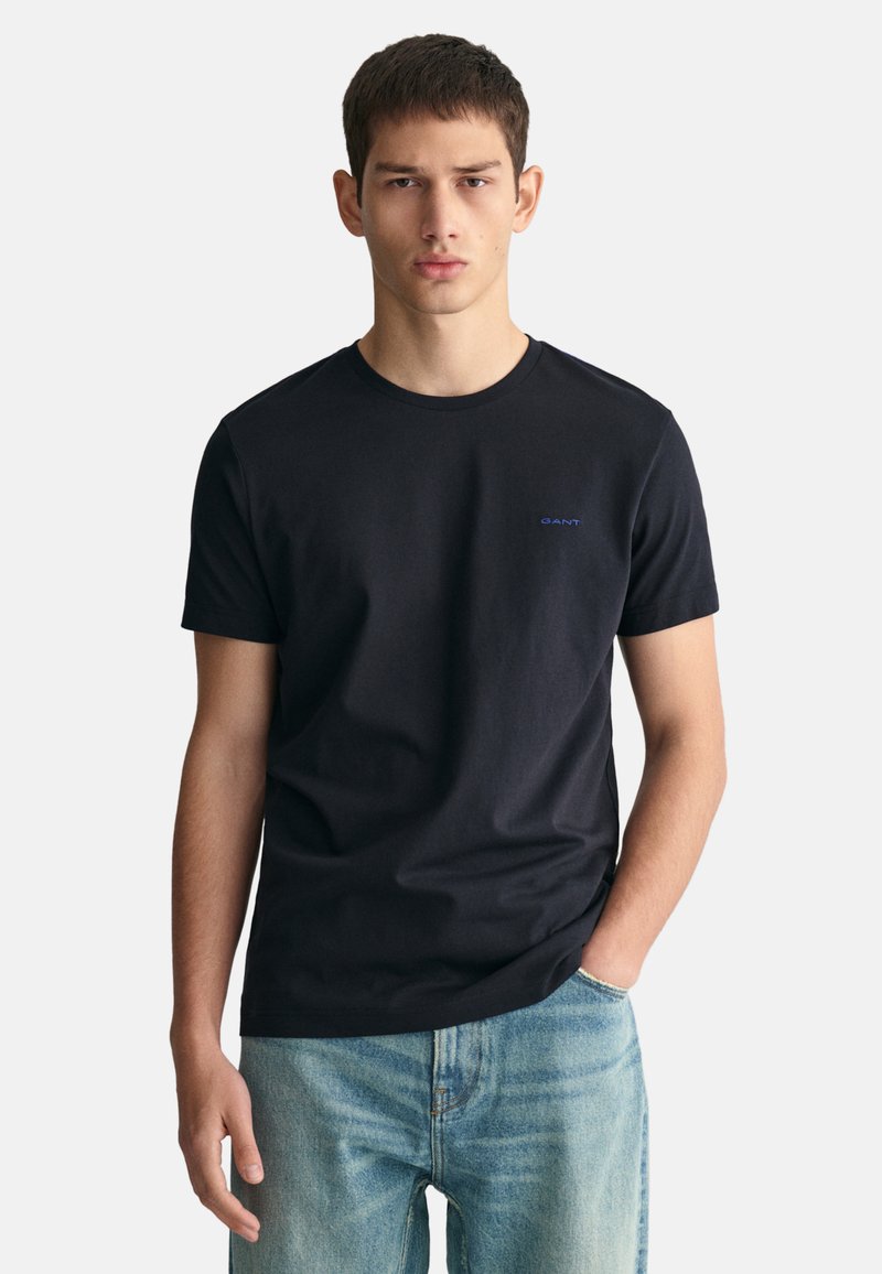 Camiseta de algodón negra con escote redondo y mangas cortas. Presenta un pequeño logo azul de "GANT" en el lado izquierdo del pecho. Combinada con unos jeans de color azul claro.