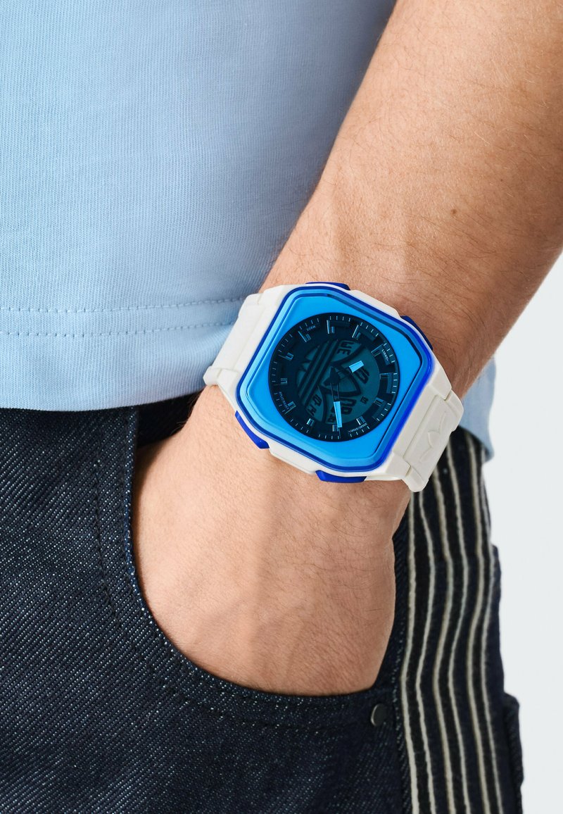 Mann trägt eine blaue und weiße analoge Armbanduhr mit quadratischem Zifferblatt, Hand in der Tasche einer dunkel gestreiften Hose, hellblaues Hemd sichtbar.