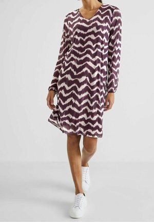 Kobieta idąca w sukience za kolano w fioletowo-biały wzór zygzaków z długimi rękawami i białych trampkach na jednolitym tle.