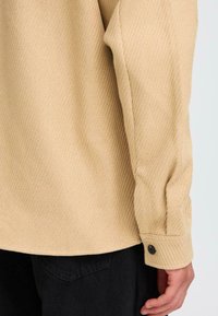 Beige textured long-sleeve shirt with a ribbed pattern, showing a buttoned cuff and part of black pants in the lower portion.