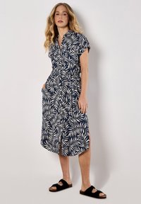 Tummansiniset mekko, jossa on valkoinen lehväkuvio, lyhyet hihat, napitus edessä, vyötärönauha ja sivutaskut. Yhdistetty mustiin sandaaleihin, polvitaipeeseen asti.