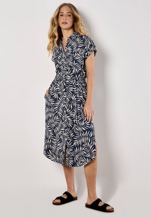Abito blu navy con motivo a foglie bianche, maniche corte, chiusura con bottoni, cintura in vita e tasche laterali. Indossato con sandali neri, lunghezza fino al polpaccio.