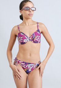 Μπικίνι με κορδόνι στο λαιμό και μοτίβο λουλουδιών σε μωβ χρώμα με ρυθμιζόμενους ιμάντες· το κάτω μέρος έχει δέσιμο στο πλάι. Το μοντέλο φοράει κομψά γυαλιά ηλίου.