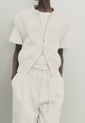 Osoba w białej koszuli z krótkim rękawem, bezrękawniku na suwak z prążkowanej dzianiny oraz wysokich białych spodniach z rękami w kieszeniach.