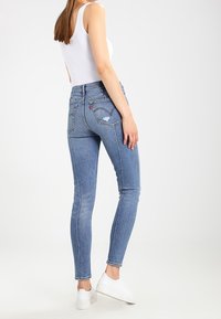 Jeans de mezclilla azul delgada con cintura alta, con bolsillos traseros y un lavado claro. Se llevan con una camiseta sin mangas blanca ajustada y zapatillas blancas.