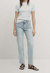 Lys grå kortærmet t-shirt, lys blå lige ben jeans og sorte sandaletter med remme. Outfittet har et simpelt, afslappet design.