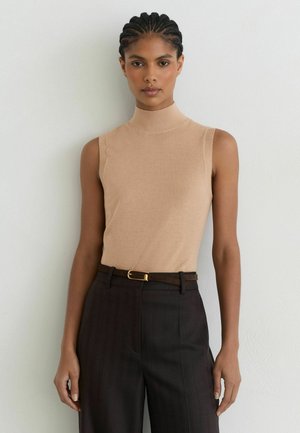 Vrouw met gevlochten haar die een mouwloos beige coltruitje draagt en donkere high-waisted broek met een dunne riem tegen een lichte muur.
