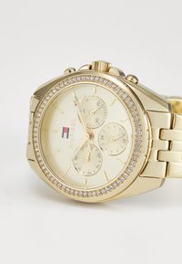 Orologio cronografo dorato con quadrante testurizzato, lunetta incastonata di cristalli, tre sottoquadranti e bracciale in metallo. Presenta un dettaglio del logo.