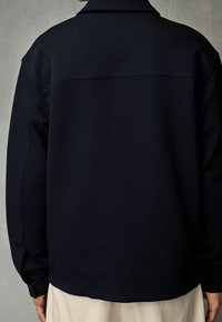 Texturovaná námořnická modrá bunda s volným střihem a manžetami na knoflíky, zdobená vzorem diamantů a čistým designem límce.