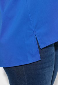 Camisa azul de manga corta hecha de tejido ligero. Presenta un dobladillo curvado con aberturas laterales y una textura suave, combinada con jeans de mezclilla oscura.