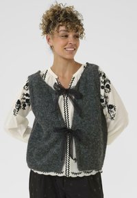 Szary, wełniany kamizelka o miękkiej fakturze, z otwartym przodem i dwoma czarnymi tasiemkami do zawiązania. Noszona na białej, haftowanej bluzce z ciemnymi akcentami.