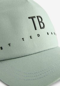 Heled roheline müts on valmistatud tekstuuriga kangast, millel on esile tõstetud must "TB" logo ja väiksemates mustades tähtedes sõnad "by TED BAKER".