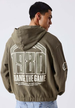 Jeune homme vu de profil portant un sweat à capuche vert olive avec un grand texte "1980 CHANGE THE GAME" dans le dos et un jean bleu.