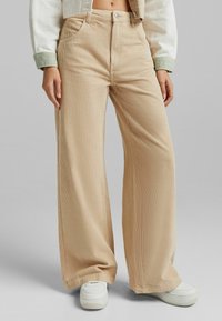 Beige korduroy kalhoty s širokými nohavicemi a vertikálními proužky, s vysokým pasem, předními kapsami a stylizované s bílými teniskami.