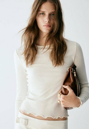 Jeune femme aux longs cheveux portant un haut côtelé crème et tenant une pochette en cuir marron sous le bras devant un fond uni.