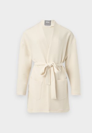 Γυναίκα που στέκεται φορώντας εκρού ζακέτα, ζιβάγκο, παντελόνια με φαρδύ πόδι και μυτερά ψηλοτάκουνα μπροστά σε λευκό φόντο.