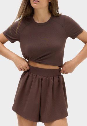 Kvinde justerer en brun cropped t-shirt og matchende højtaljede løse shorts mod en ensfarvet baggrund.