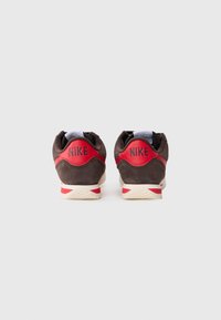 Par noge smešnih i crvenih Nike patika viđene sa stražne strane na beloj pozadini, pokazujući naziv brenda na petnom delu.