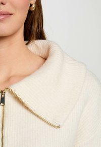 Cremefarbener Reißverschluss-Pullover mit einer ausgeprägten Rippstruktur und einem breiten, umgeschlagenen Kragen. Ausgestattet mit goldenen Reißverschlussbeschlägen und glattem Stoff.